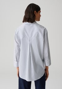 Camicia bianca con righe verticali blu navy, maniche lunghe e polsi con bottoni. Il retro presenta un design a yoke e una piega centrale per facilitare il movimento.