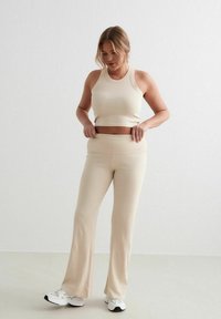 Bežely kratki top i široke hlače izrađene od rastezljivog materijala. Outfit karakteriziraju minimalni šavovi i glatka tekstura, u kombinaciji s bijelim tenisicama.