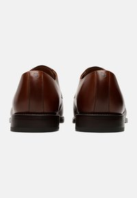 Scarpe in pelle marrone con un design elegante, tacco basso, cuciture sottili e texture liscia. Presentano una punta arrotondata e un dettaglio minimalista sul tacco.