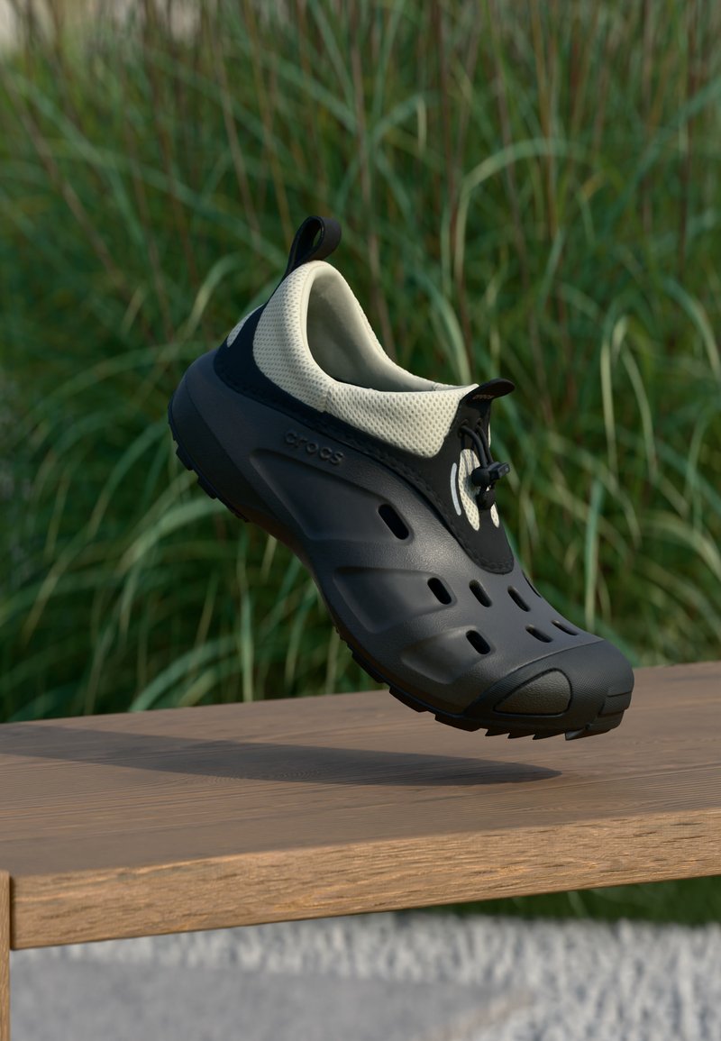 Μαύρο και μπεζ παπούτσι τύπου slip-on με οπές αερισμού και ταινίες έλξης, αιωρούμενο πάνω από ξύλινη επιφάνεια με πράσινο φόντο.