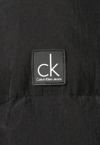 Czarna tkanina o fakturowanej powierzchni z prostokątną czarną etykietą. Na etykiecie widoczny jest biały napis "ck" oraz napis "Calvin Klein Jeans" poniżej.