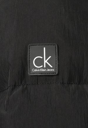 Czarna tkanina o fakturowanej powierzchni z prostokątną czarną etykietą. Na etykiecie widoczny jest biały napis "ck" oraz napis "Calvin Klein Jeans" poniżej.