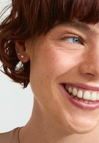 Närbild av en persons ansikte som visar två små, runda pärlörhängen. Håret är kort och rödbrunt, med synliga fräknar på huden.