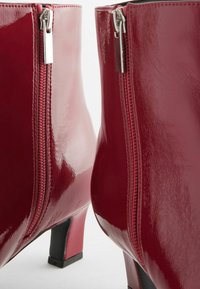 Rote Lackleder-Ankle Boots mit glänzendem Finish, silbernen Reißverschlüssen und quadratischen Absätzen; Nahaufnahme zeigt die Textur und Details.