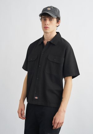 Giovane uomo che indossa una camicia nera a maniche corte con tasche sul petto e un berretto nero con un piccolo logo, in piedi davanti a uno sfondo semplice.