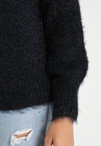 Czarny, puszysty sweter o luźnym kroju z prążkowanymi mankietami, zestawiony z przetartymi jasnoniebieskimi dżinsami. Tekstura wydaje się miękka i futrzasta.