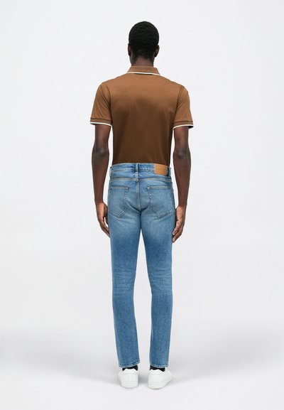 Camisa polo marrón con ribete blanco, vaqueros azules de corte recto y zapatillas blancas, vistas desde atrás contra un fondo liso.