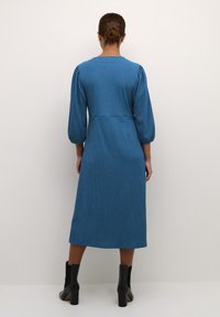 Robe midi bleue texturée avec manches bouffantes et taille cintrée. Portée avec des bottines noires. Encolure ronde simple sans motifs visibles.