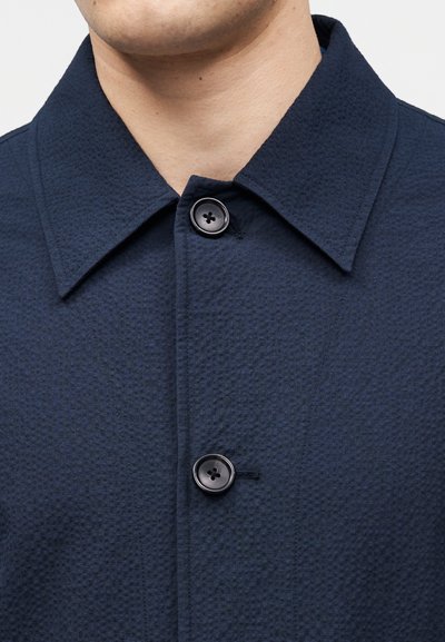 Άνδρας που φοράει υφασμάτινο μπλε σκούρο πουκάμισο με γιακά, κουμπωμένο στον λαιμό και το στήθος, ορατός από το πηγούνι μέχρι το επάνω μέρος του στήθους.