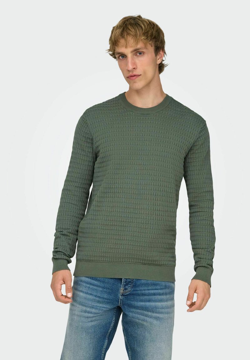 Pull vert texturé à col rond, manches longues et poignets côtelés. Porté avec un jean bleu clair.