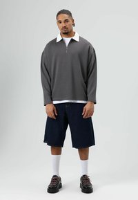 Grijze sweatvest met rits en een witte kraag, gecombineerd met donkerblauwe denim shorts. Het model draagt witte sokken en zwarte schoenen met roze accenten.