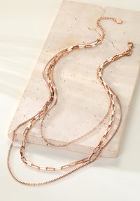 Trójwarstwowy naszyjnik w kolorze różowego złota z różnymi wzorami łańcuchów: płaskie ogniwa, skręcana linka i gładkie wykończenia. Ustawiony na tle jasnego kamienia.
