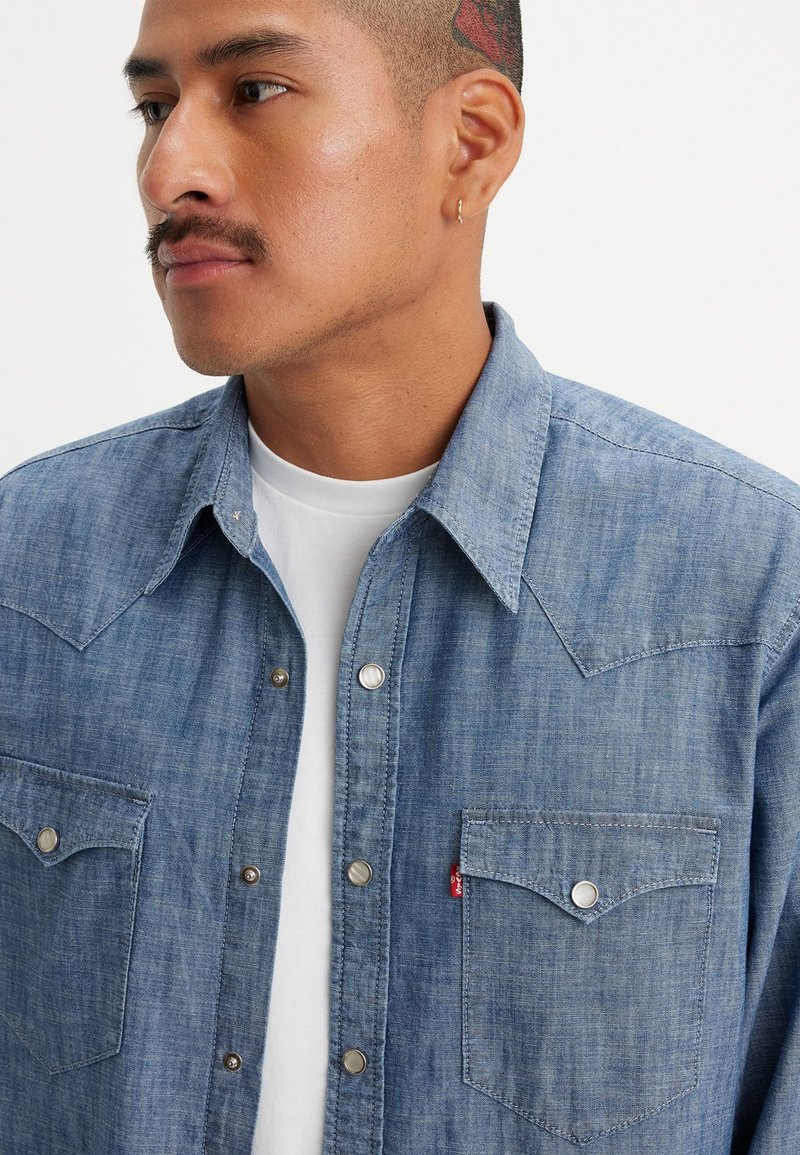 Chemise en denim bleu clair avec des boutons-pression, un design de patte pointue, deux poches poitrine et une petite étiquette logo sur la poche.