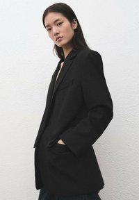 Blazer negro con un diseño estructurado, solapas con muesca y dos bolsillos frontales. La tela parece lisa y tiene un ajuste entallado.