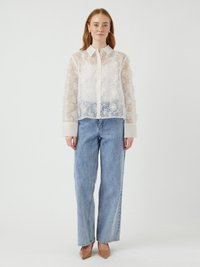 Blusa in pizzo floreale bianco trasparente con chiusura a bottoni, abbinata a jeans in denim azzurro chiaro a vita alta e scarpe con tacco beige a punta.