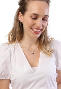 Zilveren ketting met een hanger in de vorm van een bloem, gedragen boven een witte V-hals top met pofmouwen. Gladde stof textuur, lichte kleur.