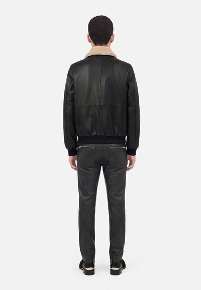 Homme debout de dos, portant une veste en cuir noire avec un col en fourrure beige et un pantalon en cuir noir assorti avec des poches zippées.
