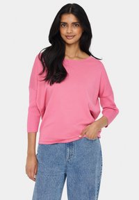 Brīvi pieguļoša, ribota rozā jakā ar trīs ceturtdaļu piedurknēm, apaļu kakla izgriezumu, valkāta ar augsto jostu zilām džinsām. Gluda tekstūra, ikdienišķs stils.