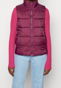 Plommonfärgad pufferväst med hög krage och framdragkedja, med quiltat mönster. Bärs över en långärmad rosa topp och kombineras med ljusblå jeans.