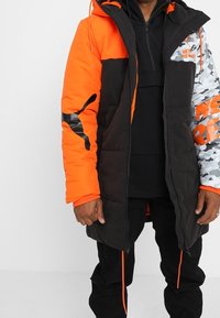 Giacca imbottita con un design a blocchi di colore nero e arancione, con dettagli in camouflage e un logo. Include un cappuccio e un orlo regolabile.