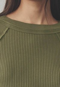 Pull côtelé vert olive avec un large décolleté et un tissu texturé. Détail de surpiqûres visibles le long des épaules.
