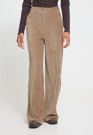 Persoon die beige corduroy broek met hoge taille en wijde pijpen draagt, een donkerbruine top met lange mouwen en bruine schoenen, staand tegen een witte achtergrond.