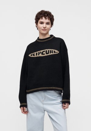 Jeune personne aux cheveux courts et bouclés portant un pull noir Ripcurl et un jean bleu clair sur un fond uni.