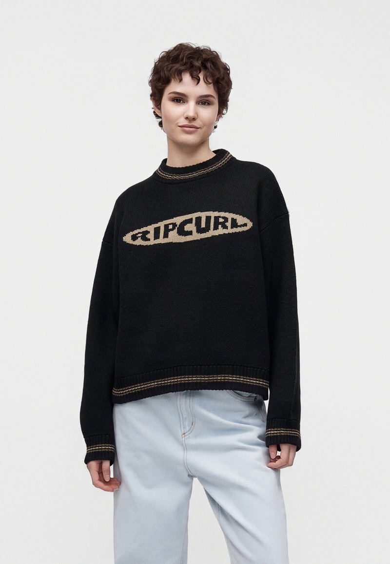Jeune personne aux cheveux courts et bouclés portant un pull noir Ripcurl et un jean bleu clair sur un fond uni.
