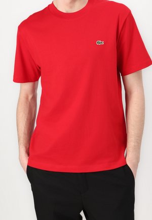 Homme portant un t-shirt rouge uni à manches courtes avec un petit logo vert de crocodile sur la poitrine gauche et un pantalon noir.
