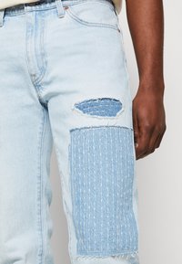 Ljusblå denimjeans med ett slitaget knä som har en lapp av texturerad tyg, vertikal sömnad och fransade kanter.