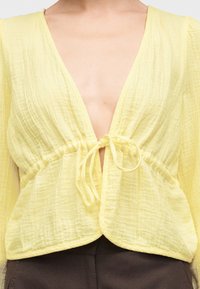 Gros plan d'une personne portant un chemisier jaune clair texturé à col en V profond et noué à l'avant, associé à un pantalon foncé.