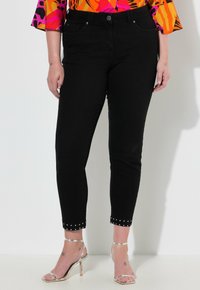 Černé džíny z denimu s přiléhavým střihem, zkrácenou délkou a cvočkovým detailem na manžetách, doplněné stříbrnými sandály na podpatku.