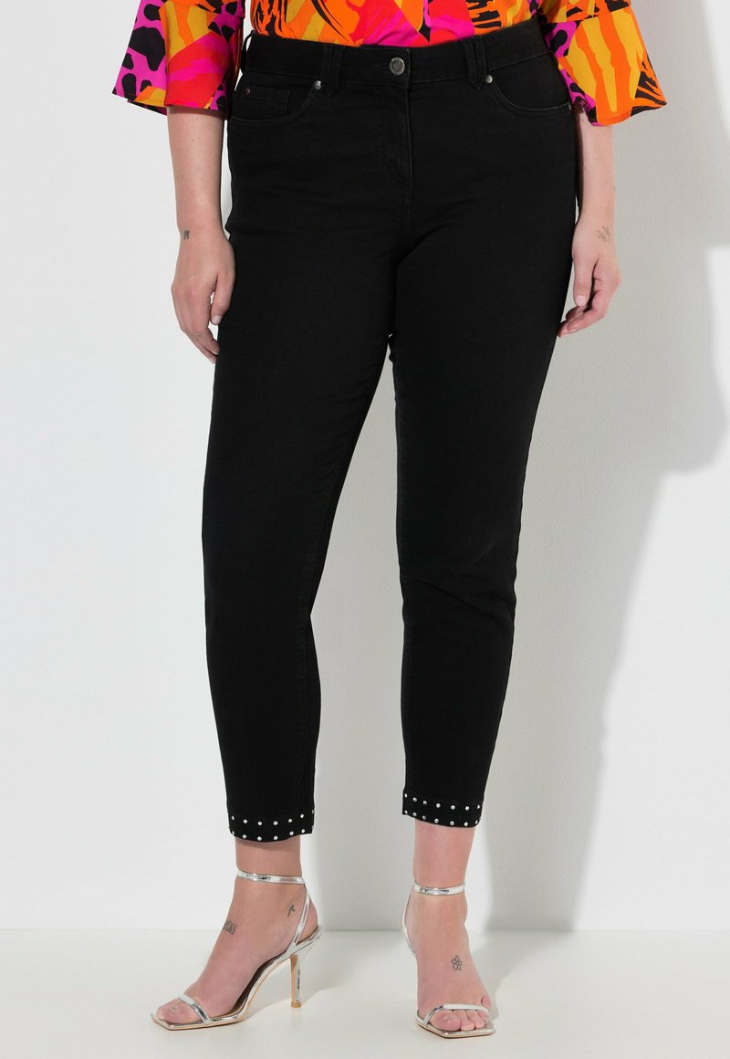Černé džíny z denimu s přiléhavým střihem, zkrácenou délkou a cvočkovým detailem na manžetách, doplněné stříbrnými sandály na podpatku.