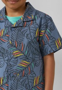 Kortekorte overhemd van lichtblauwe stof met verschillende bladerenpatronen in groen, geel, rood en oranje, met twee knopen aan de voorkant.