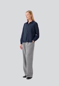 Camicia blu navy con colletto, abbinata a pantaloni larghi in lana grigio chiaro. Scarpe nere, in piedi contro uno sfondo semplice.