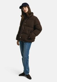 Bruine pufferjack met een hoge kraag, drukknopen en twee zijzakken, gecombineerd met blauwe jeans en een marineblauwe baseballcap.