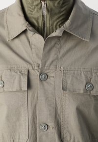 Veste marron clair boutonnée sur pull vert côtelé à fermeture éclair, montrant les détails du col et de la poche poitrine.