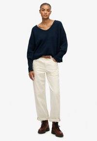 Oversized granatowy sweter dzianinowy z szerokim dekoltem w kształcie litery V, w połączeniu z białymi spodniami o prostym kroju i brązowymi butami na sznurówki. Lekka tekstura.