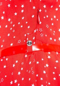 Roter Stoff mit weißem Sternmuster, versehen mit einem Reißverschluss, einem kleinen silbernen Knopf mit der Aufschrift "KS" und einer glänzenden roten Schlaufe, die durch den Stoff gefädelt ist.