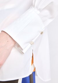 Camisa blanca de manga larga con detalle de puños plisados y acentos de botones dorados. Tejido ligero; diseño minimalista con una textura suave.