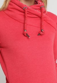 Suéter de color coral con un alto cuello tipo cowl, que presenta cordones texturizados y acentos metálicos, fabricado en un suave tejido texturizado.