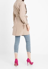 Beżowy płaszcz trencz z paskiem w talii i wiązanymi rękawami, zestawiony z podartymi jasnoniebieskimi dżinsami i różowymi szpilkami. Gładka faktura i dopasowany krój.
