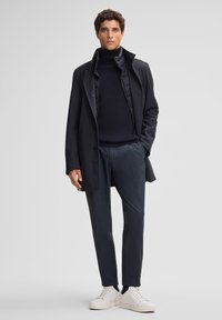 Czarny płaszcz zewnętrzny, prążkowany czarny golf i fakturowane ciemne spodnie. Białe sneakersy. Gładkie tkaniny, dopasowany krój, brak widocznych wzorów.