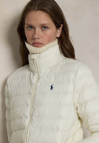 Jonge vrouw met lang bruin haar die een witte gewatteerde jas draagt met een hoge kraag en een klein donkerblauw polo-logo op de borst.