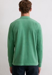 Mann trägt ein langärmliges grünes Polohemd und dunkle Hosen, steht vor einer weißen strukturierten Wand.