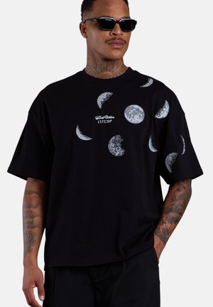 Mann trägt schwarze Sonnenbrille und schwarzes übergroßes T-Shirt mit mehreren Mondphasen-Motiven und tätowierten Armen.