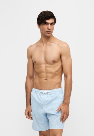 Shirtless man with short dark hair wearing light blue and white striped swim shorts standing against a plain light background.