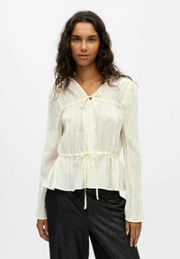 Blouse à manches longues crème avec un ourlet peplum, fermeture à nouer au col, tissu texturé et léger éclat, associée à un pantalon noir brillant.