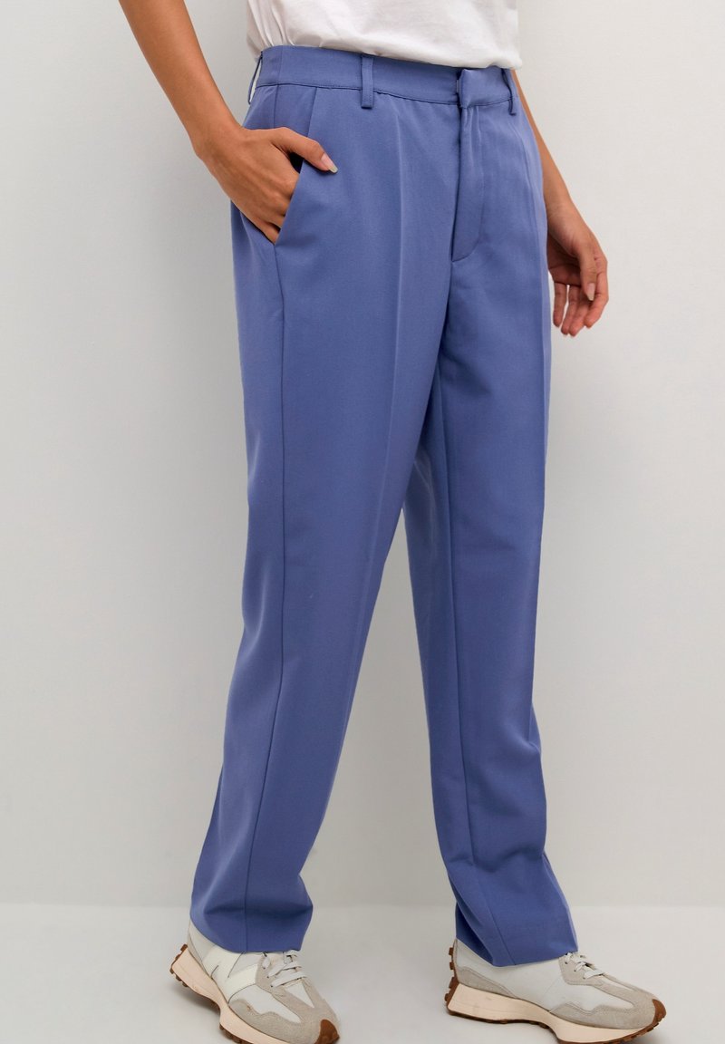 Un pantalon bleu sur mesure présente un tissu lisse, une coupe droite, des poches latérales et une taille mi-haute, associé à des baskets blanches et beiges.
