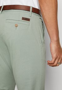 Lichtgroene broek met een gladde textuur, voorzien van een bruine leren riem, een achterzak met een knoop en een merklabel.